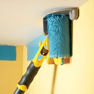 A blue paint roller is being used to paint a yellow wall, positioned against a ceiling. The roller is attached to a yellow extendable handle.