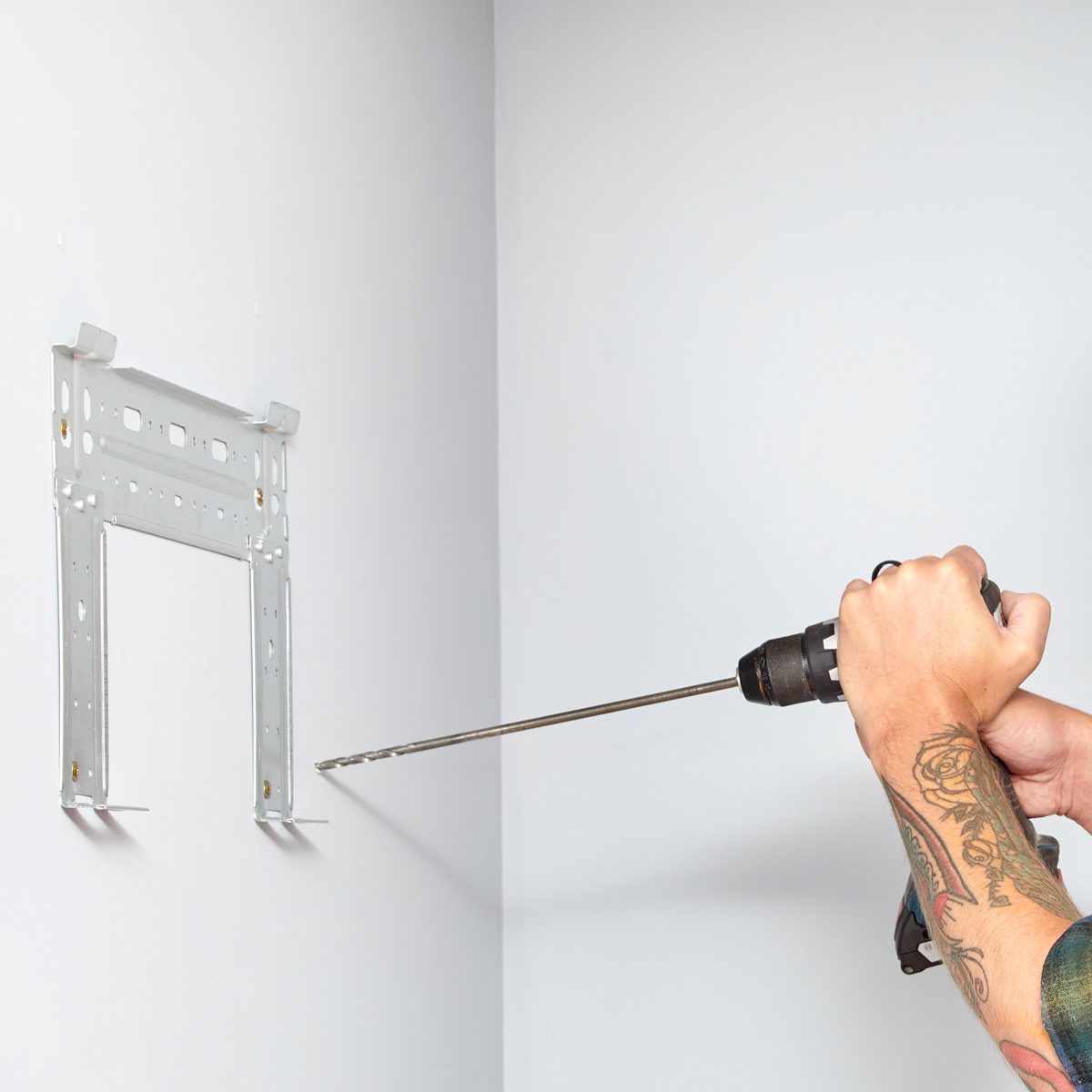 A hand holds a power drill, drilling into a wall where a metal bracket is mounted, in a plain, well-lit room.