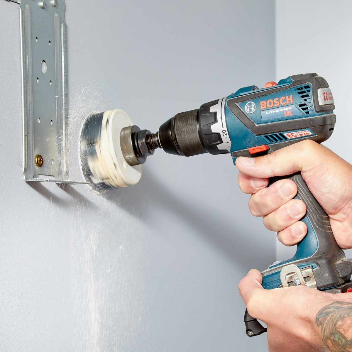 A person uses a power drill on a wall, creating dust, while holding the tool against a mounting bracket. The wall is painted light gray.