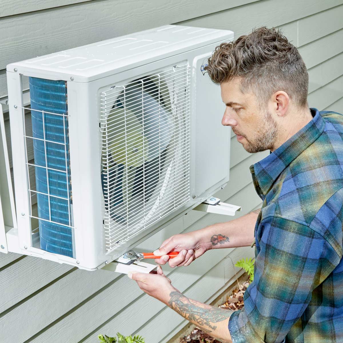A man is adjusting an outdoor air conditioning unit using a wrench, positioned beside a house with wooden siding and some foliage at the base.