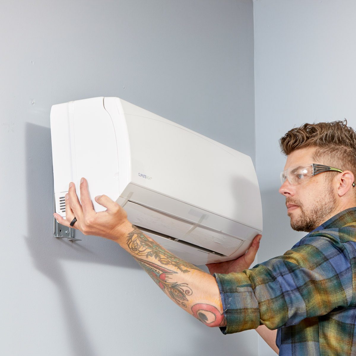 A man installs a wall-mounted air conditioner, using both hands to secure it while wearing protective glasses, in a room with light blue walls.
