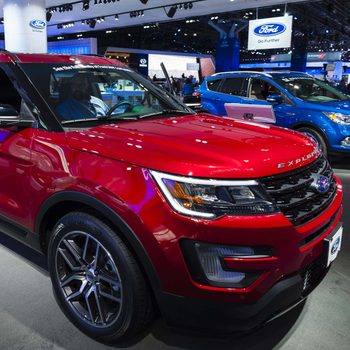 A red Ford Explorer is showcased at an auto show, with a person seated inside. Other vehicles and display booths are visible in the background.