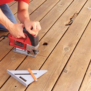A person uses a jigsaw to cut wooden planks on a floor, with a square tool and pencil nearby for measurement.