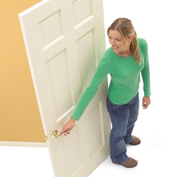 A woman in a green shirt opens a white door with a brass handle, smiling, in a simple indoor setting with a beige wall.
