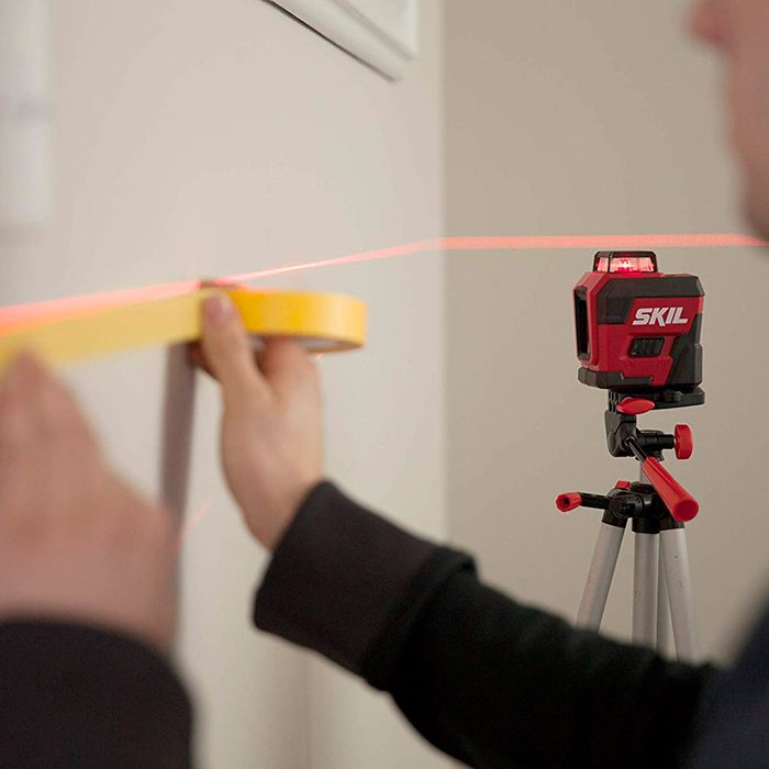A person holds yellow tape along a wall while aligning it with a red laser line projected from a tripod-mounted laser level.