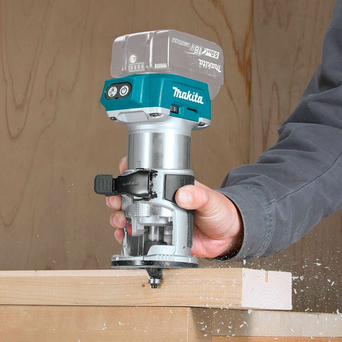 A hand holds a Makita router, cutting through a wooden plank, while wood shavings fly in the air against a wooden background.