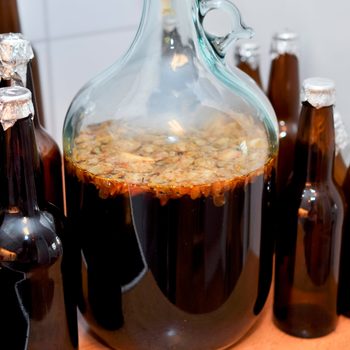 A glass carboy contains dark liquid with visible sediment, surrounded by several brown bottles, suggesting a brewing or fermentation process in a kitchen setting.