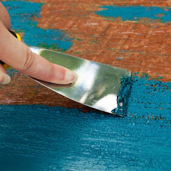 A hand holds a metal scraper, smoothing turquoise paint on a wooden surface, with remnants of different shades visible in the background.
