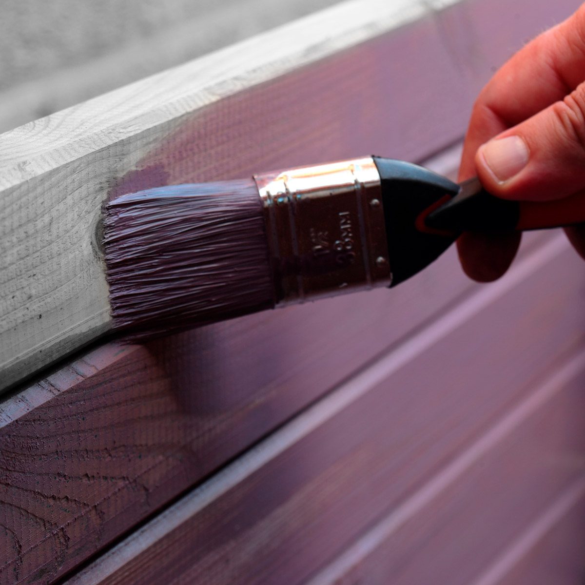 A paintbrush applies purple paint to wooden planks, while a hand steadies it in a light, neutral setting.