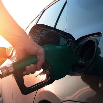 A hand holds a green fuel nozzle, inserting it into a car's gas tank. The background features sunlight, suggesting a late afternoon setting.