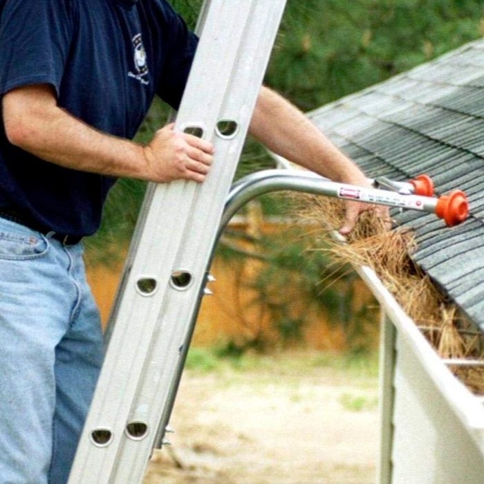 A man on a ladder is cleaning debris from a gutter using a specialized tool in a residential outdoor setting surrounded by trees.