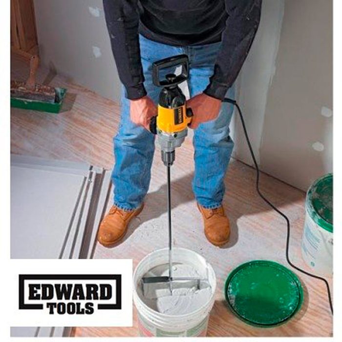A person uses a power mixer to blend materials in a bucket on the floor, surrounded by construction tools and materials in a room.