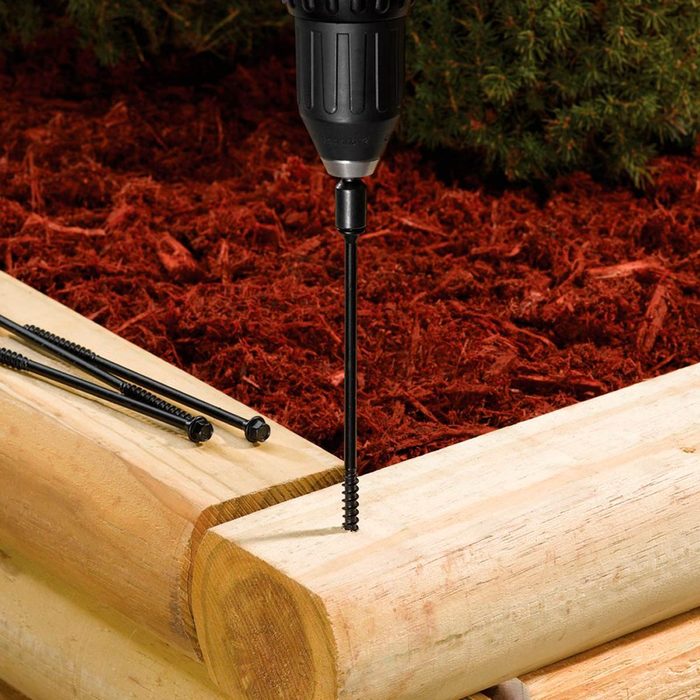 A drill is securing a black screw into a wooden log, surrounded by red mulch and additional black screws nearby.
