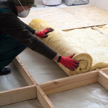 A person in red gloves rolls out insulation material on a wooden framework, preparing for construction in a possibly unfinished indoor space.