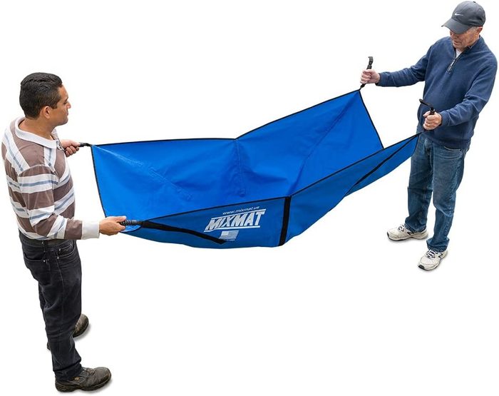 Two men hold a large blue fabric mat, stretching it between them. They are outdoors, preparing for use, possibly for a camping or outdoor activity.