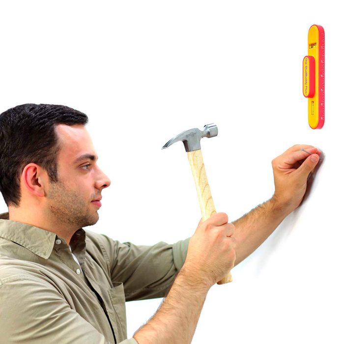 A man holds a hammer poised to strike a nail into a white wall while a level tool is mounted nearby for guidance.