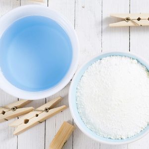 A bowl of blue liquid and a bowl of white powder sit on a white wooden table, accompanied by scattered wooden clothespins.