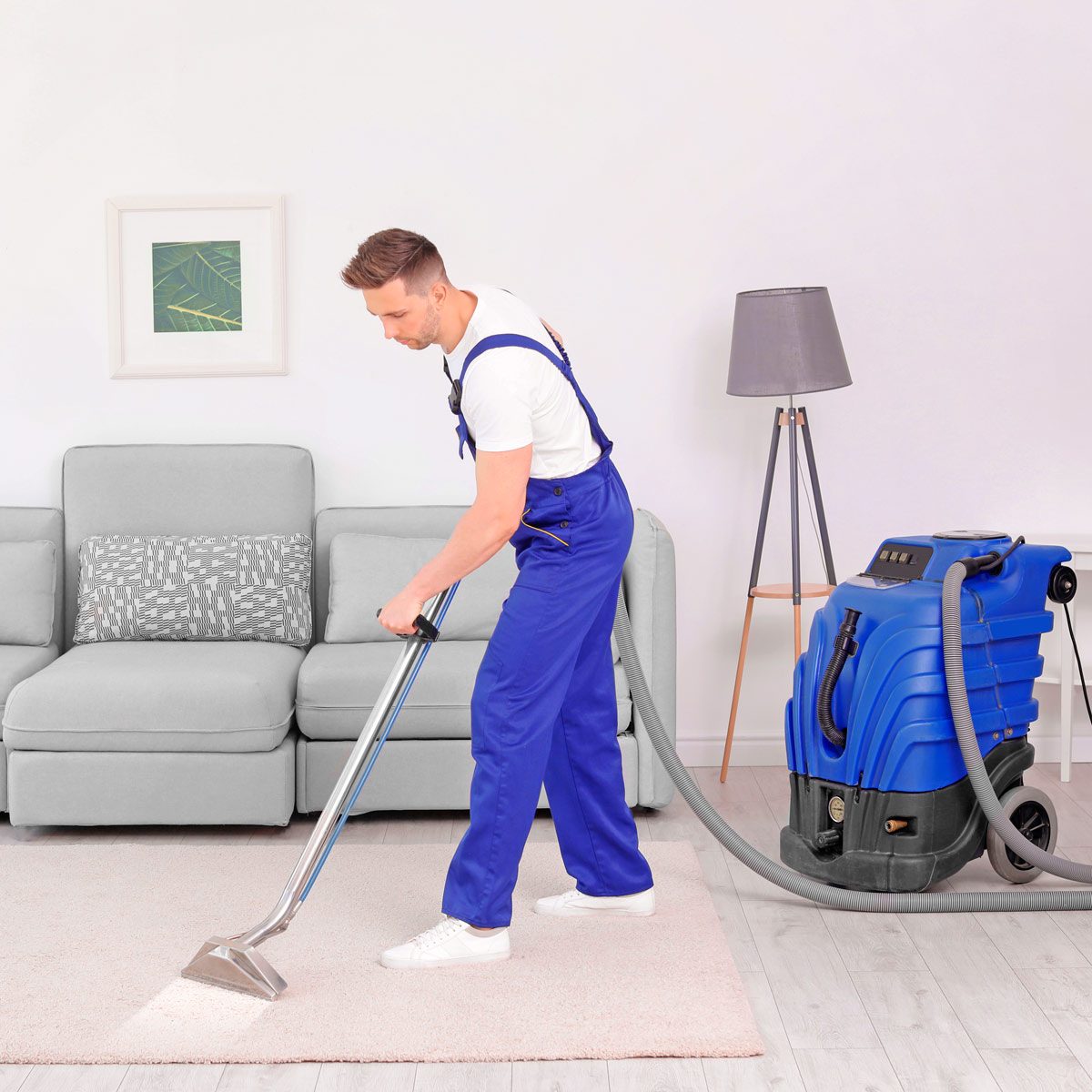 A man in blue overalls vacuums a light carpet with a machine in a modern living room featuring a gray sofa and a lamp.