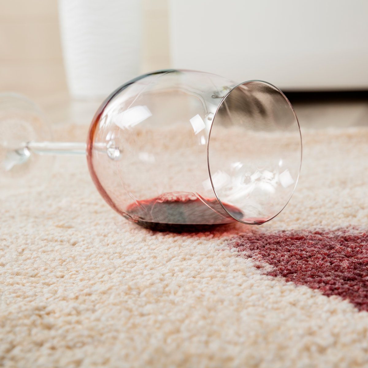 A wine glass lies on its side, spilling red wine onto a cream-colored carpet, creating a stark contrast in the cozy indoor setting.