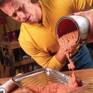 A man in a yellow shirt pours pink paint from a can into a metal tray, spilling some in a workshop with tools in the background.