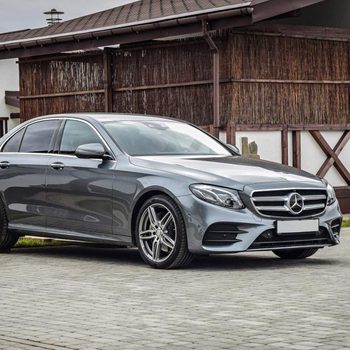 A sleek gray car is parked on cobblestone near a rustic building with a thatched roof and wooden accents, under an overcast sky.