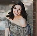 A young woman smiles brightly while leaning against a brick wall, showcasing a relaxed style in a light off-the-shoulder top, with a blurred background.