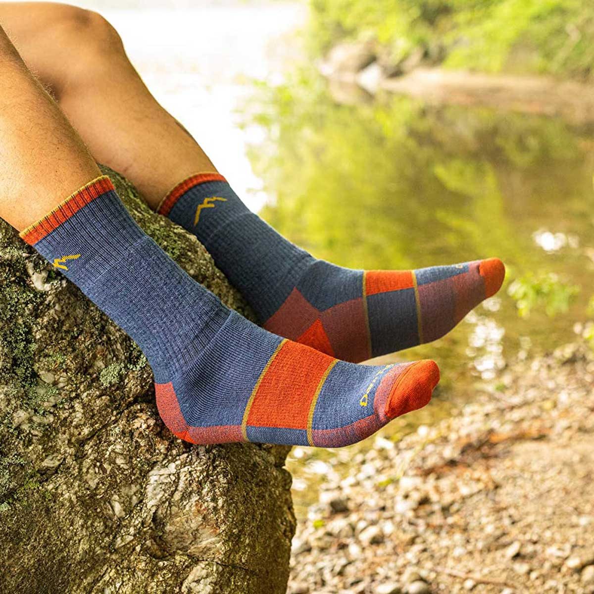 Colorful socks rest on a rock, as a person relaxes by a stream surrounded by lush greenery. Sunlight filters through the trees.