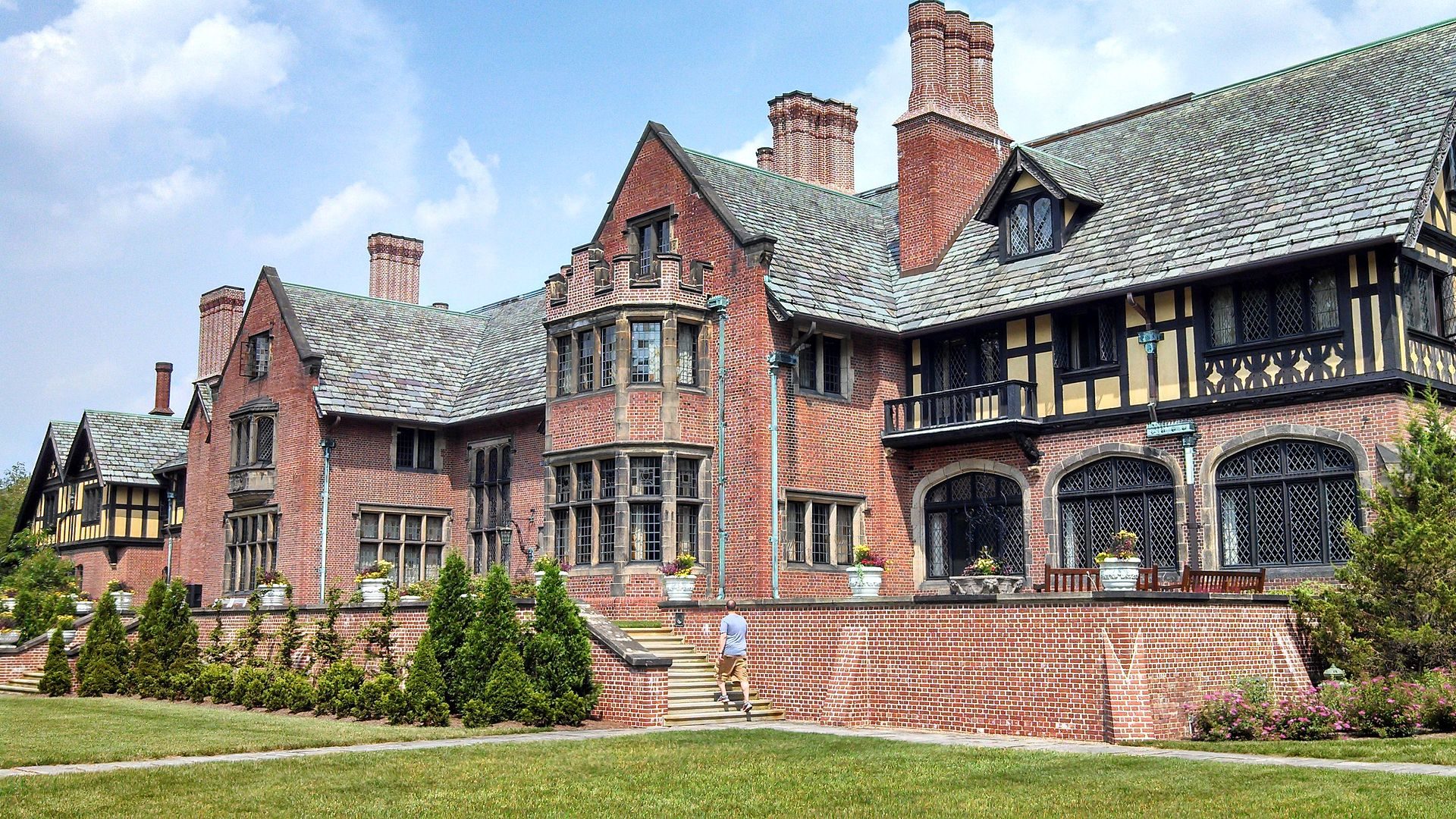 A large brick mansion with ornate windows stands in a landscaped yard, while a person walks up stone steps towards the entrance on a sunny day.