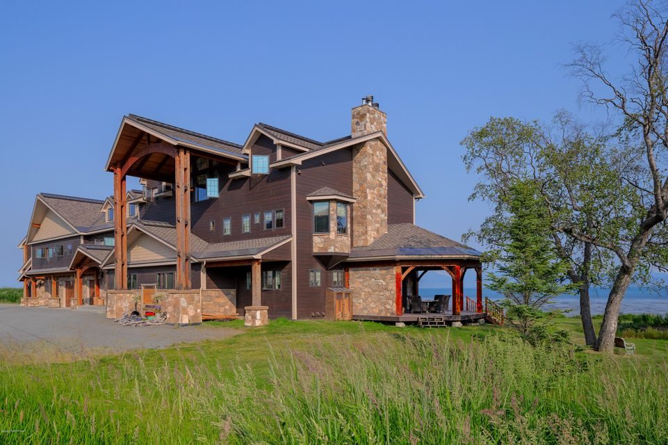 A large, modern house with wooden pillars and stone accents stands amidst green grass, facing a body of water under a clear blue sky.