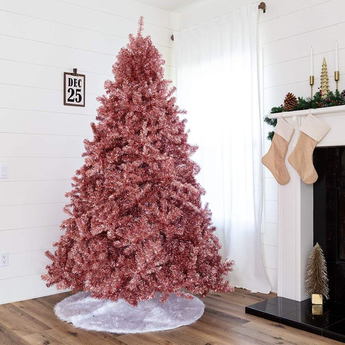 A pink Christmas tree stands decorated in a living room, surrounded by a white tree skirt, with stockings hanging by a fireplace and a calendar showing December 25.