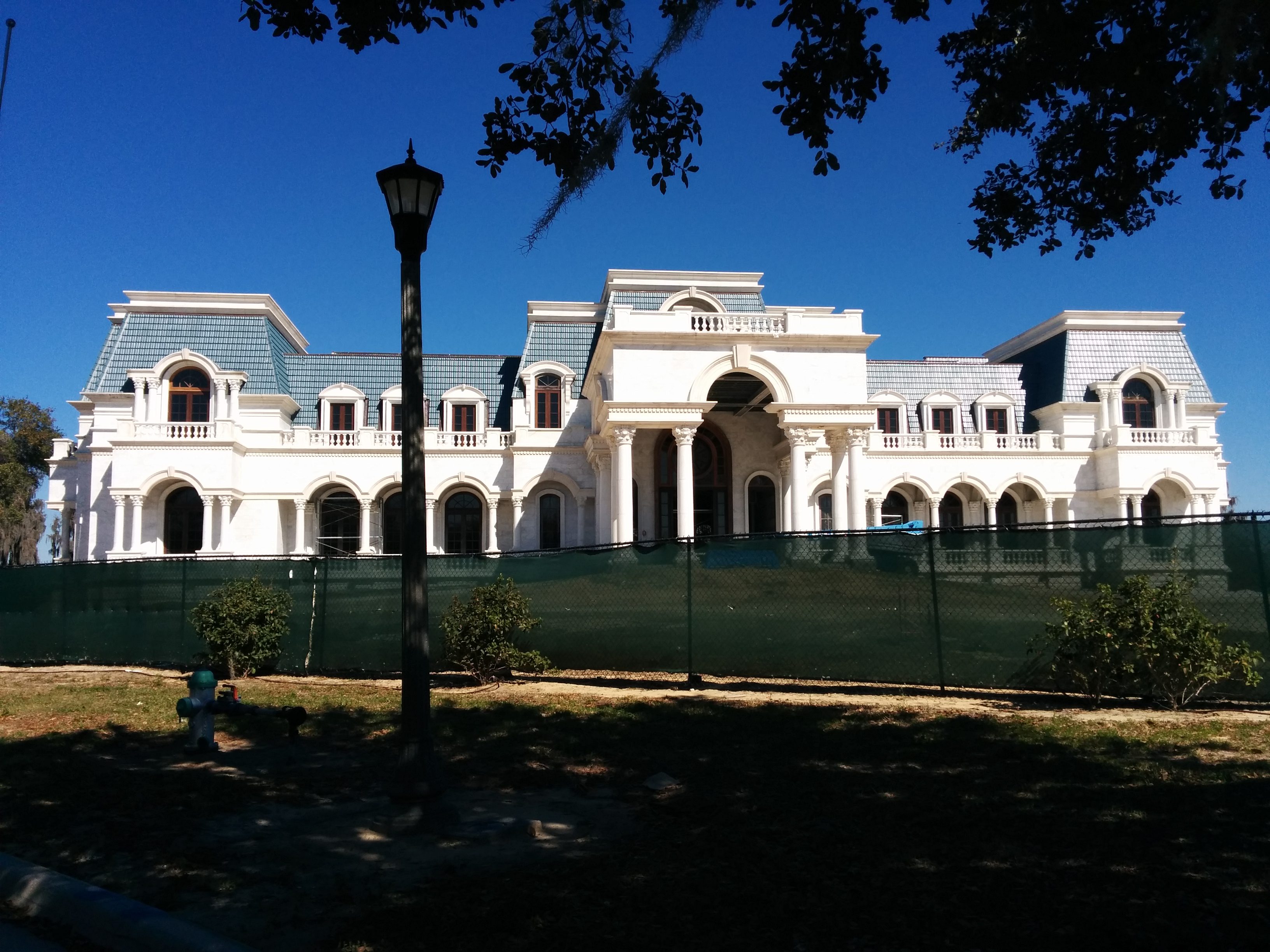 A large, white mansion with multiple gables stands behind a green construction fence under a clear blue sky, surrounded by grassy land and trees.