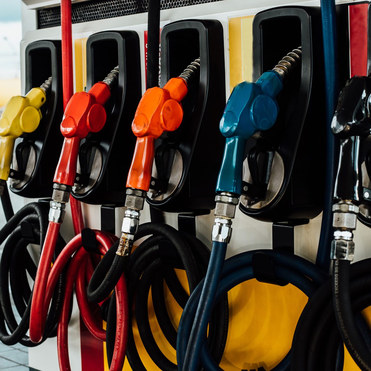 Gasoline pump nozzles in various colors hang from a dispenser, indicating different fuel types available at a gas station, with hoses coiled below.