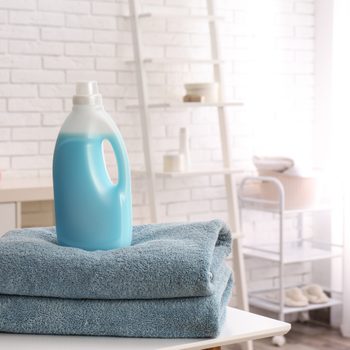 A blue detergent bottle rests on two stacked blue towels in a bright, airy room with white brick walls and soft textiles in the background.