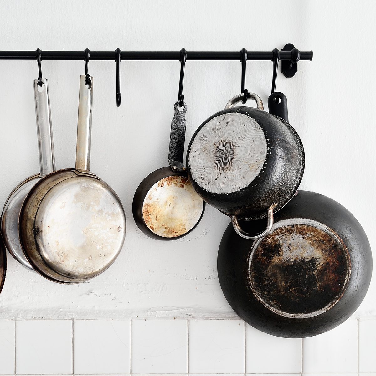 Pans and pots hang from a black rod against a white wall, showcasing various sizes and degrees of wear from cooking.