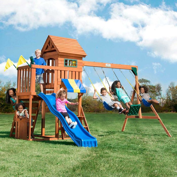 A wooden playset with a slide and swings is occupied by children playing joyfully on a grassy field under a blue sky with clouds.