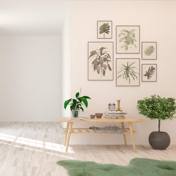 A wooden table holds plants, decor, and books in a bright room with light wood flooring. Green plants and framed botanical prints decorate the walls.