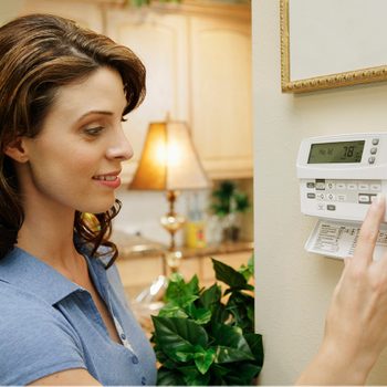 A woman adjusts a digital thermostat on a wall in a well-lit kitchen, surrounded by plants and a lamp in the background.