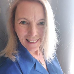 A woman with long, light brown hair smiles, wearing a blue shirt, set against a softly lit background with delicate, swirling patterns.