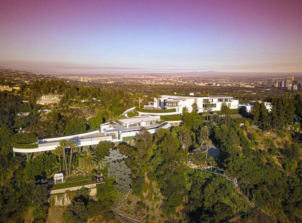 A modern mansion is perched on a hillside, surrounded by trees, showcasing a sprawling view of a city skyline in the distance under a clear sky.