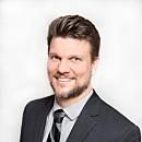 A man in a suit smiles confidently, standing against a plain white background, projecting a professional and approachable demeanor.