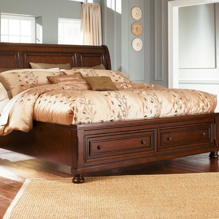 A wooden bed with a plush, decorated comforter rests in a softly lit room, surrounded by light-colored walls and a woven rug beneath it.