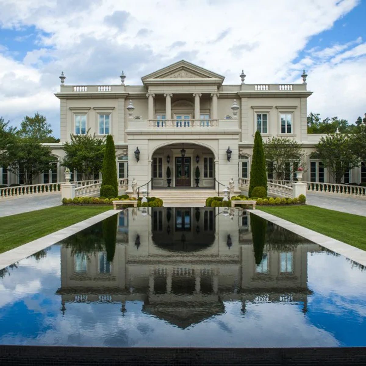 A grand mansion stands prominently, reflecting in a calm pool, surrounded by manicured lawns and lush greenery under a partly cloudy sky.