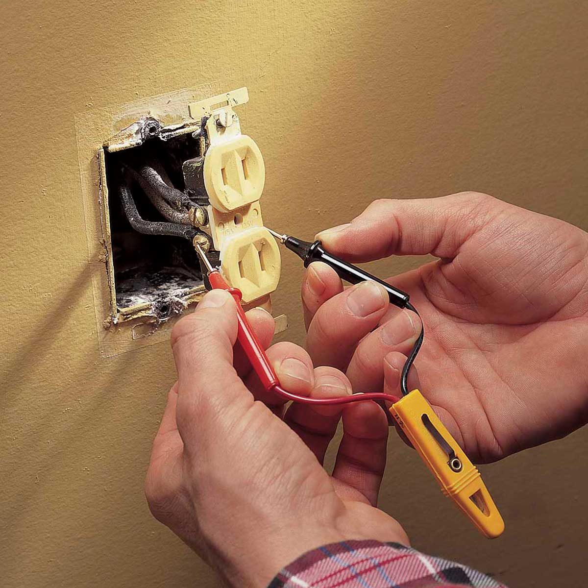 A person uses a multimeter with red and black leads to test electrical outlets mounted on a wall, surrounded by yellow paint and wiring.