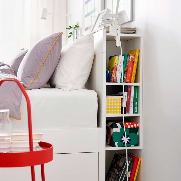 A white bedside shelf holds books and decorative boxes, next to a bed with soft pillows. A red side table features a glass and stacked books.