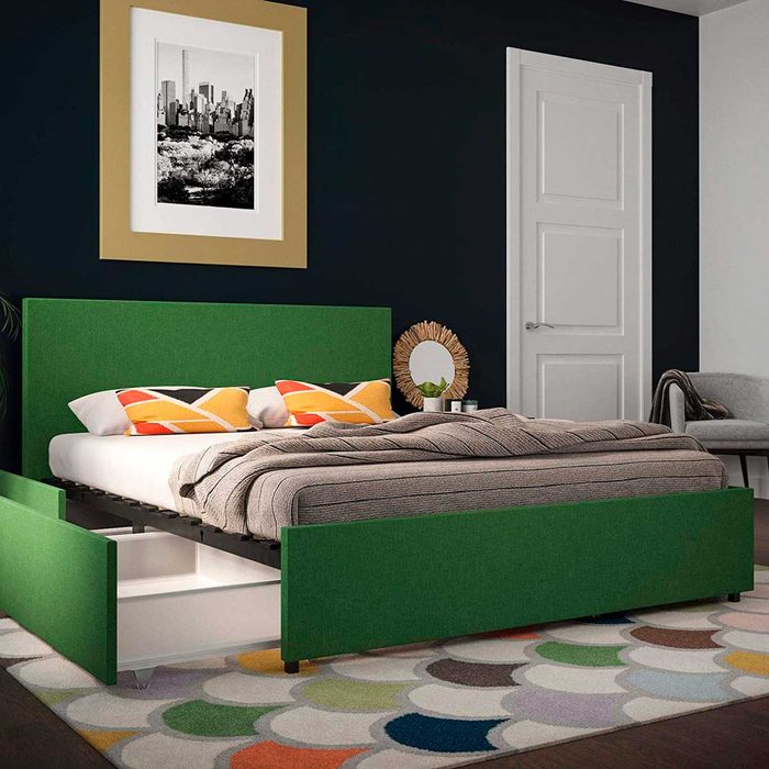A green upholstered bed with patterned pillows and a cozy blanket occupies a room featuring a colorful rug, dark walls, and a wall-mounted artwork.