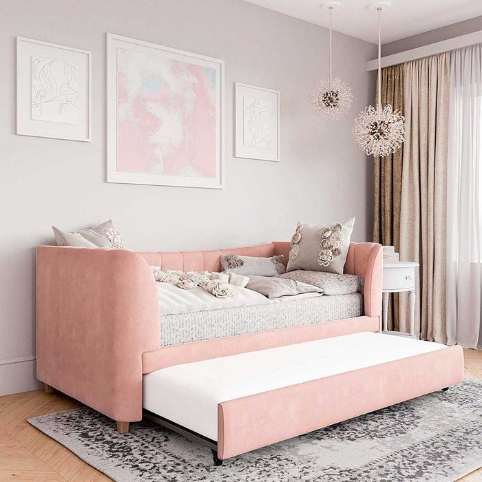 A pink daybed with decorative pillows is positioned against a wall, while a white pull-out mattress rests below, in a softly lit, elegantly decorated room.