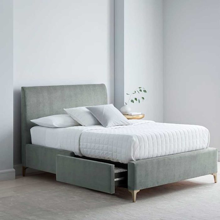 A soft, green upholstered bed with a white quilted cover is positioned in a minimalist room, featuring a bedside table and plant.