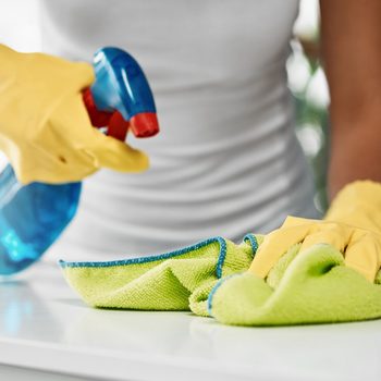 A person in yellow gloves sprays a cleaning solution onto a green cloth while preparing to clean a surface in a bright environment.