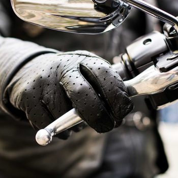 A gloved hand grips a motorcycle's throttle, poised for acceleration against a blurred urban background. The leather glove features perforations for breathability.