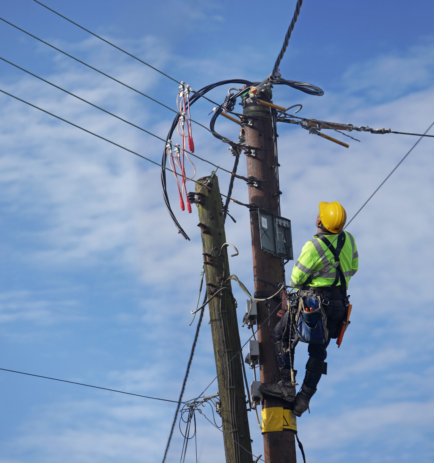 A worker, wearing a hard hat and reflective clothing, climbs a utility pole to perform maintenance on overhead electrical wires against a clear blue sky.